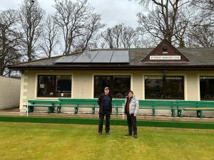 Two members of the St Ninian Bowling Club standing on the bowling green. The newly installed solar panels on the roof of the club house are visible in the background.